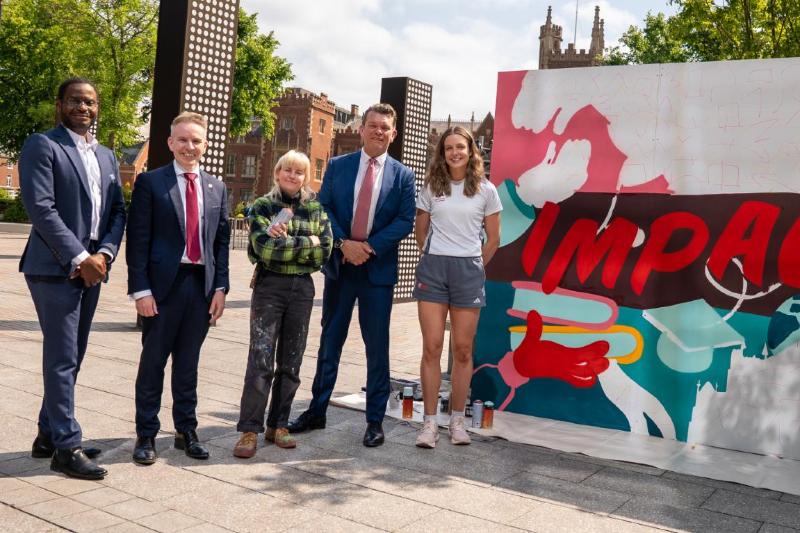 Two women and three men stand posed beside a piece of art which reads the word Impact
