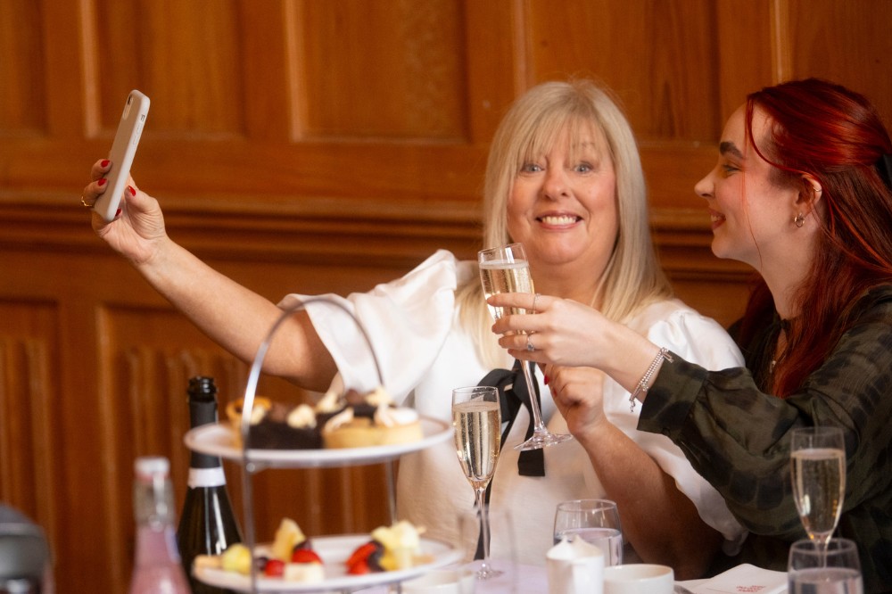 two women seated at a table with afternoon tea - the lady on the left is holding her phone up for a selfie while looking to the camera and smiling