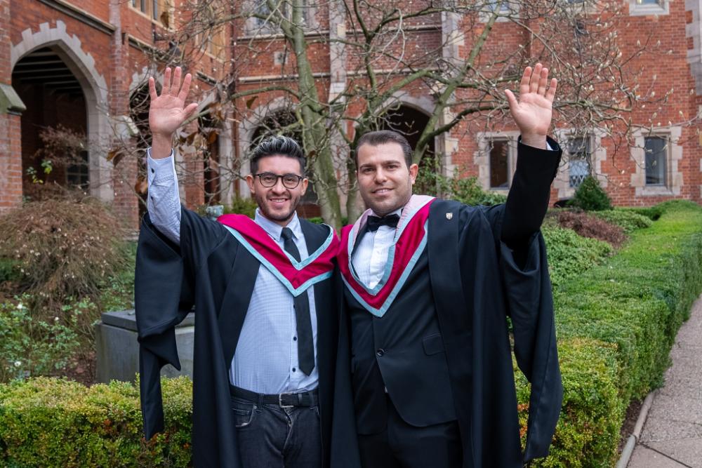 Two male graduates smiling and raising their hands in celebration outside a Queen's University Belfast building.