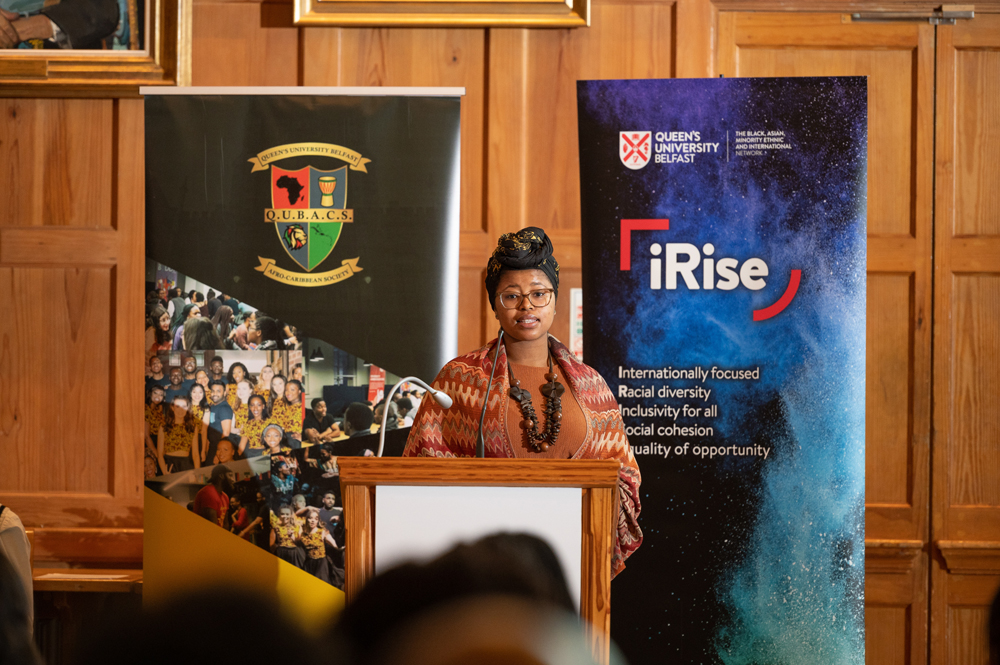 Student speaking from podium in Great Hall, showing QUBACS and iRise banners in background