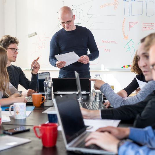 A group of staff sitting around a table at a team meeting while a manager stands loooking at a document