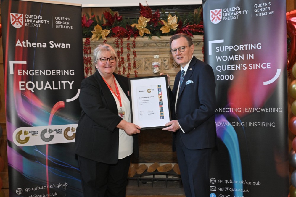 woman pictured with a man in a suit holding a framed award certificate in an old wood-paneled event hall with large fireplace and presentation screen in the background and pop-up stands and balloon pillars on either side