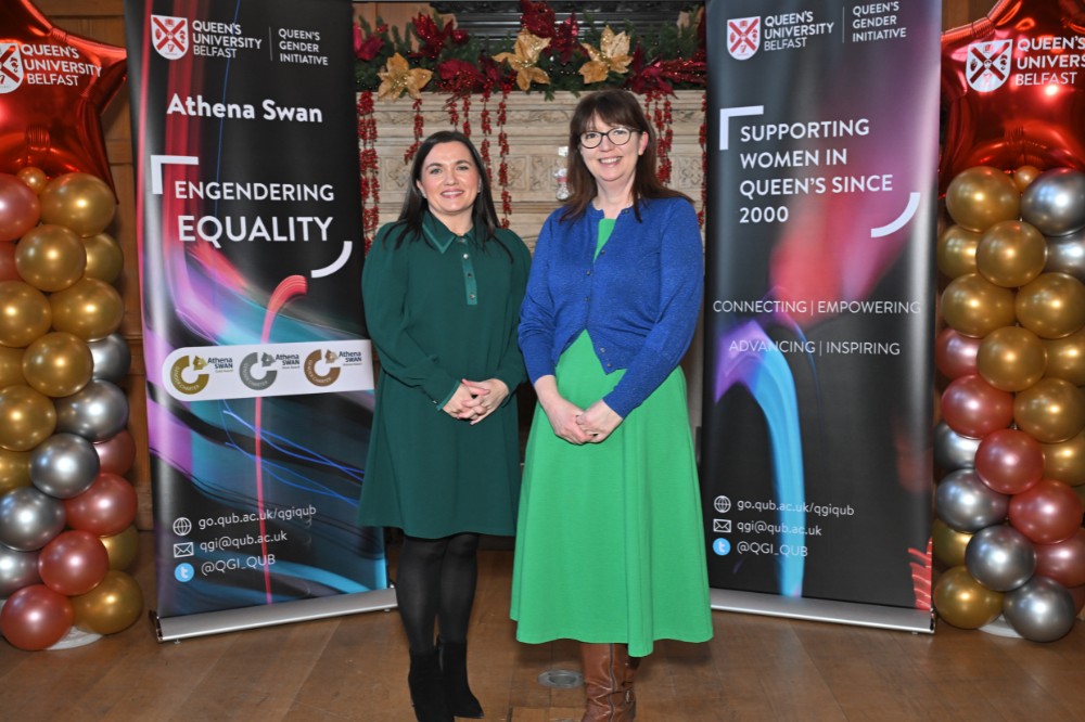 two women posing for a photo in an old wood-paneled event hall with pop-up stands and presentation screen in the background