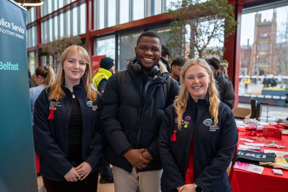 Two Queen’s Students’ Union representatives stand smiling beside an attendee at an information stall during a campus event.