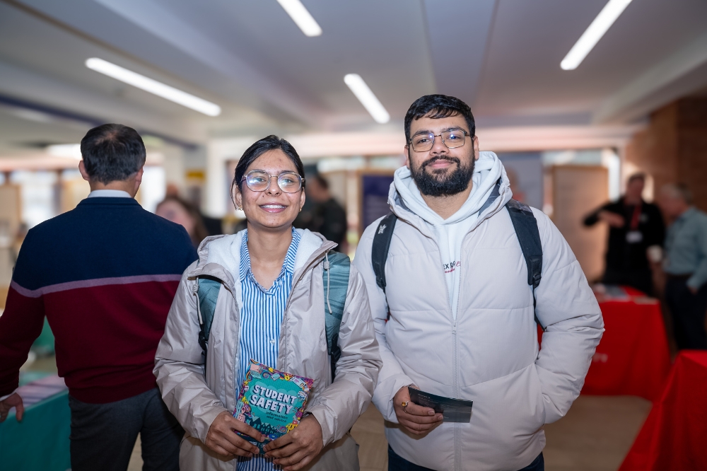 Two students stand smiling at an indoor campus event, holding information leaflets, with event stalls visible in the background