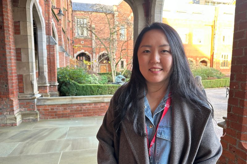 A woman with long dark hair stands smiling in an outdoor cloister area, with red-brick university buildings and gardens in the background