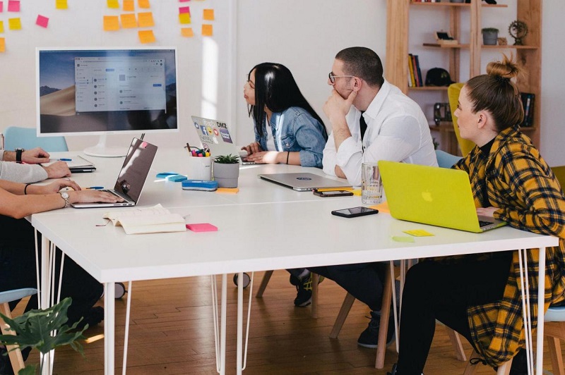 five people with laptops and work accessories sitting at an informal meeting table looking at a mounted screen