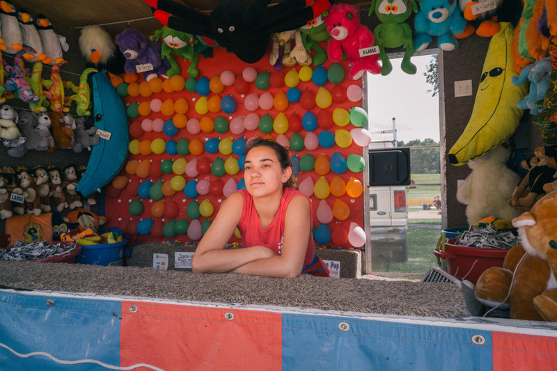 photo exhibit by American photographer Andrew White, showing young woman standing in a fairground booth selling soft toys and balloons