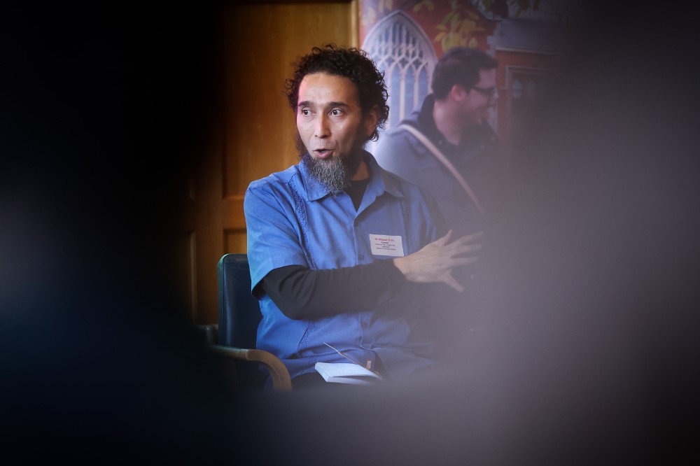 seated man speaking at an indoor event, with blurred elements in the immediate foreground