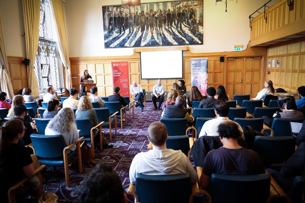 view from the back of a comfortable-looking, brightly lit, wood-paneled, indoor meeting space, showing group of seated people listening to a speaker at a podium and seated panel. A large painting hangs on the back wall.