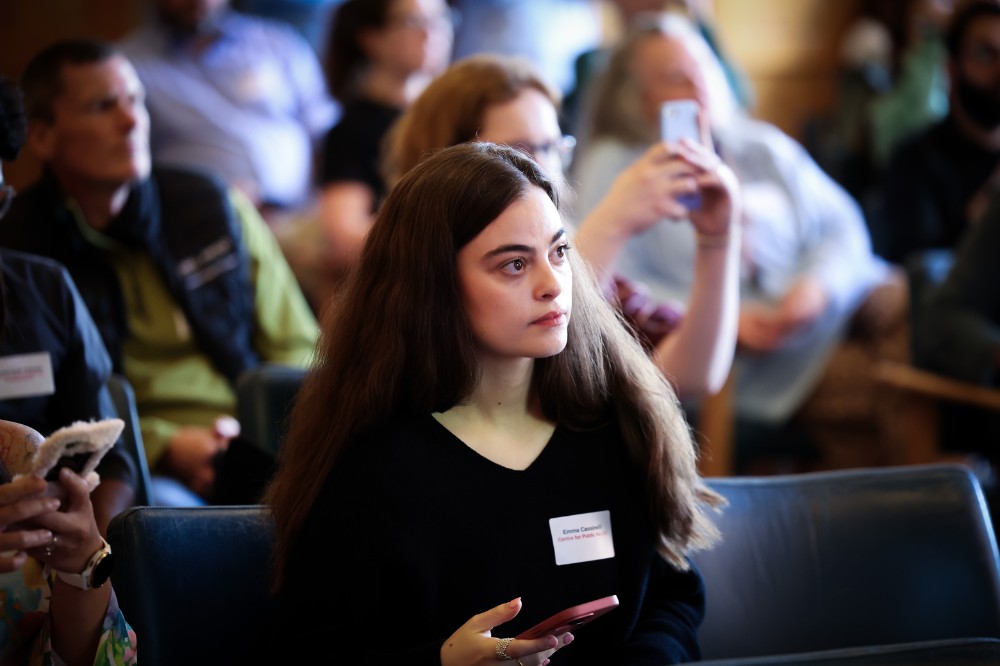 seated woman with long, dark hair listening to a presentation at an indoor event, with other attendees pictured in the background