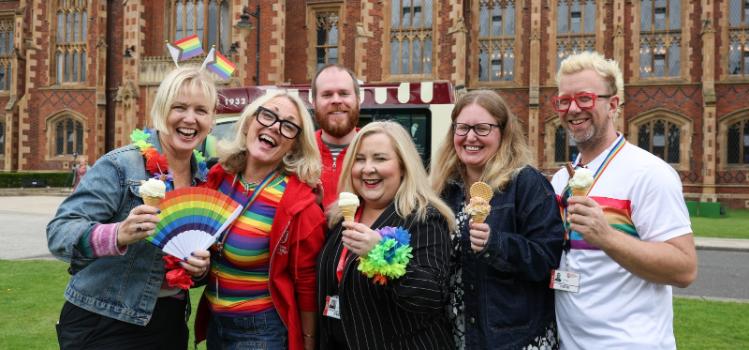 group of smiling people wearing rainbow paraphernalia in front of a red-brick building