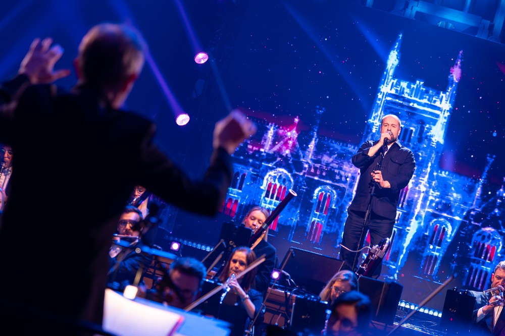 man singing on a blue-lit stage with an orchestra and conductor pictured in the mid and foreground