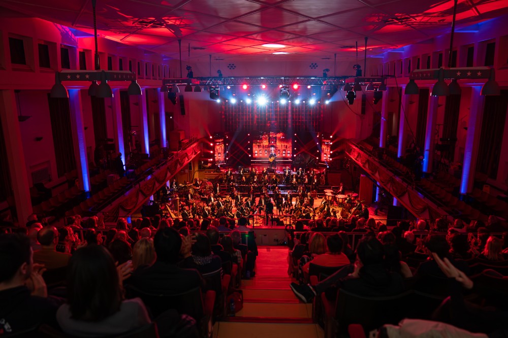 long view from the back of the balcony of a large hall showing a seated audience in the foreground and seated orchestra on stage in the background against a presentation screen