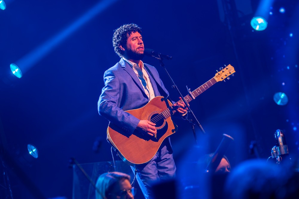 singer-songwriter wearing a suit and playing a guitar on a blue-lit stage