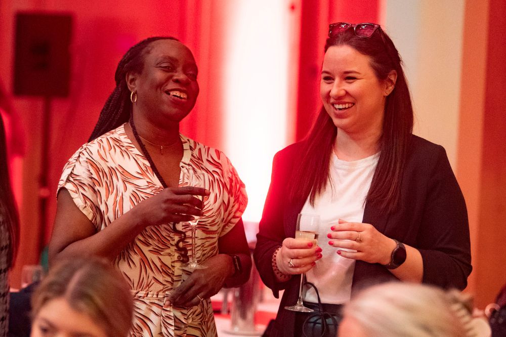 two women holding drinks in champagne flutes sharing a light moment at an indoor event