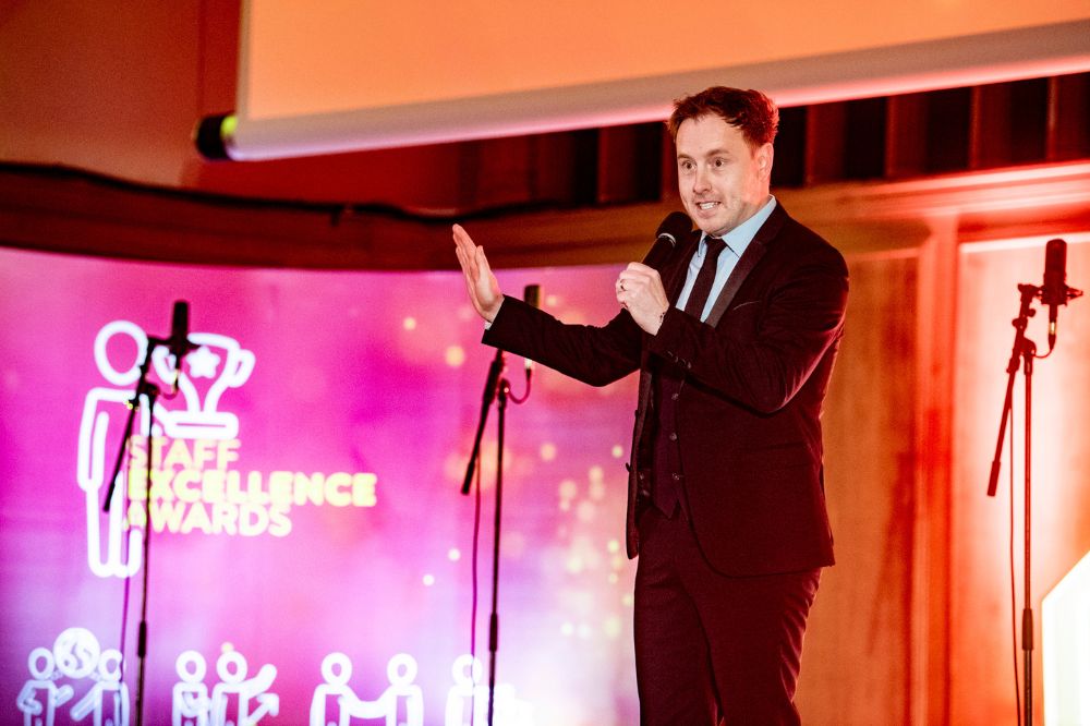 Man in suit speaking into a microphone at an event. A pop-up stand to his right reads 'Staff Excellence Awards'.
