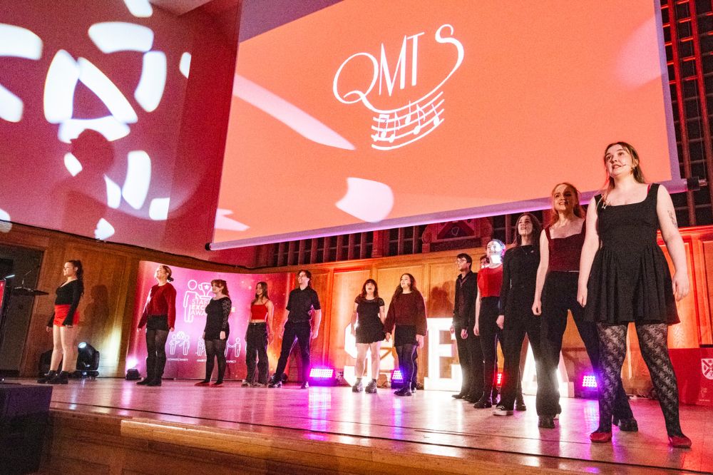 group of young women and men performing on a stage with a large presentation screen behind them at an indoor event