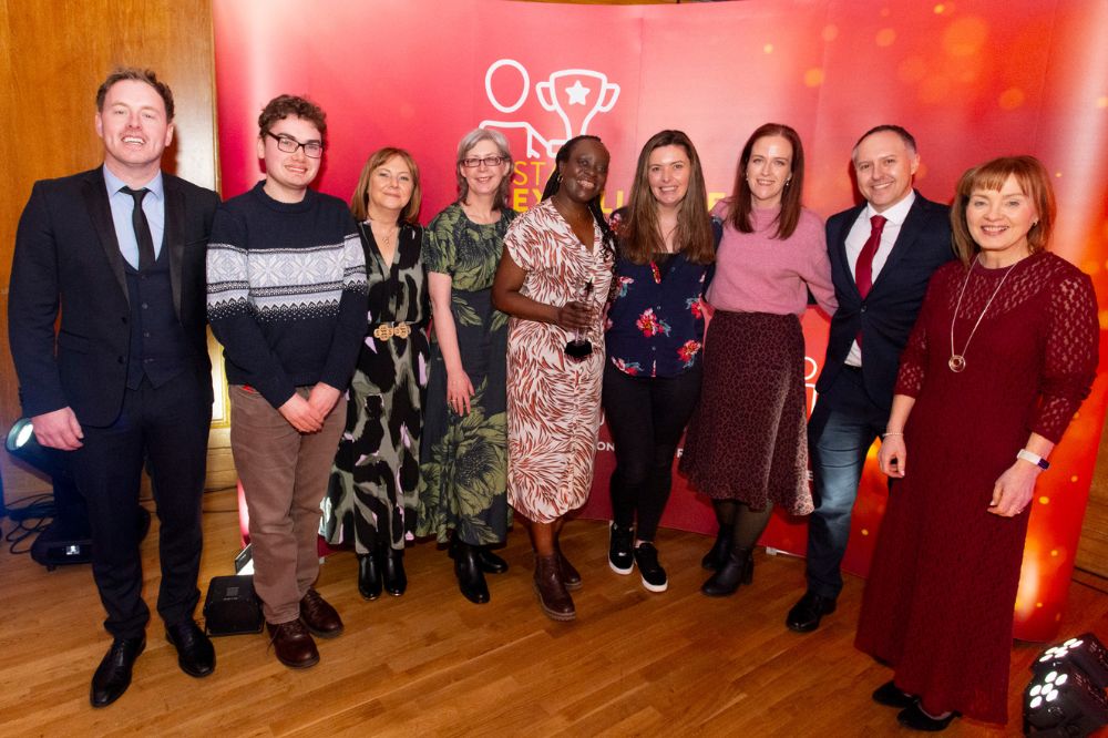 Group of three men and six women posing for the camera at an indoor awards event. The woman in the centre is holding a crystal trophy.