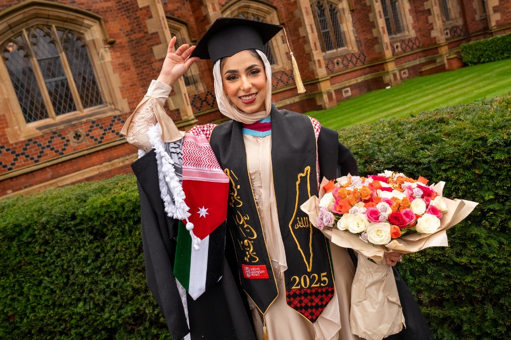 woman wearing graduation robe, hat and scarf with Arabic lettering, holding a bouquet of flowers in front of an old building