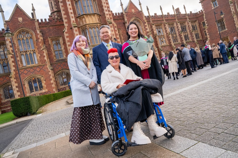 two women and one man standing with a woman in a wheelchair - all dressed in formal clothing - with an old redbrick building and tower in the background